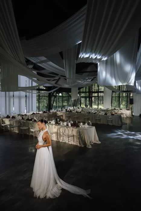 Bride in white gown gazing down in a dimly lit reception hall adorned with draped fabric and candles.