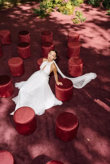 Bride in flowing white gown seated among scattered red velvet stools in an outdoor wedding space.