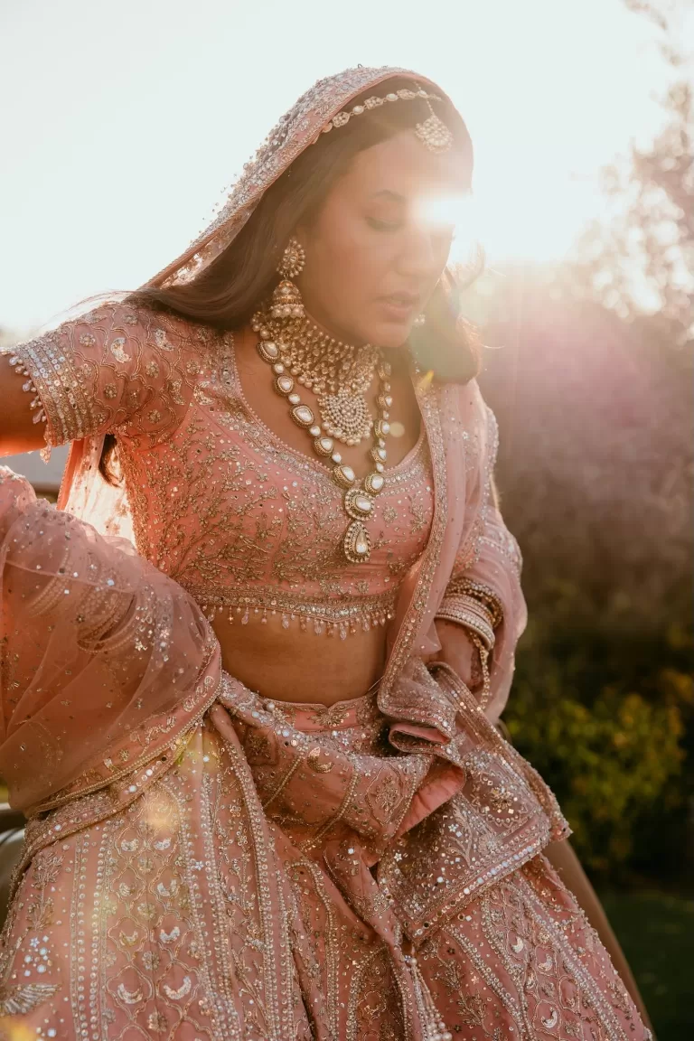 Bride in beaded pink lehenga at golden hour, sunlight streaming over Cigarral de las Mercedes gardens.