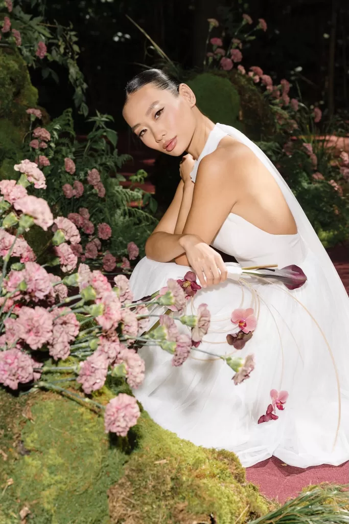Bride holding orchids and calla lilies while seated among moss and pink flowers in a woodland setting.