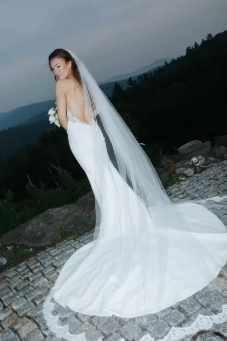 Bride looking over her shoulder in a sleek gown and long veil, standing on a stone path with mountain views at Château Ste. Agnès.