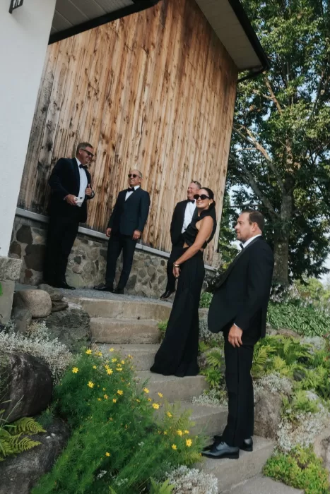 Elegant wedding guests arriving at Château Ste. Agnès, dressed in black tie against a rustic wood and stone exterior.