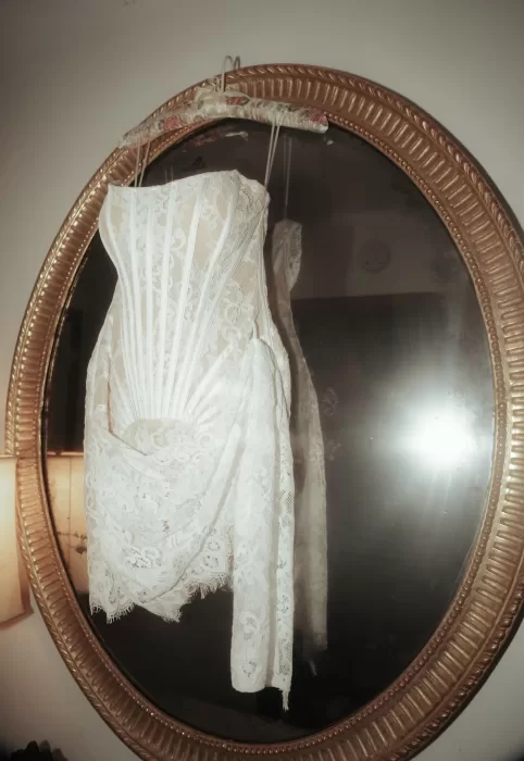 Bride’s lace mini dress hanging from a floral hanger on a gold oval mirror at Château Ste. Agnès.