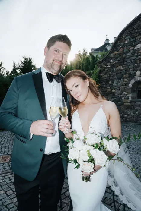 Newlyweds toasting with champagne at Château Ste. Agnès, standing on cobblestones with stone walls and turrets behind them.