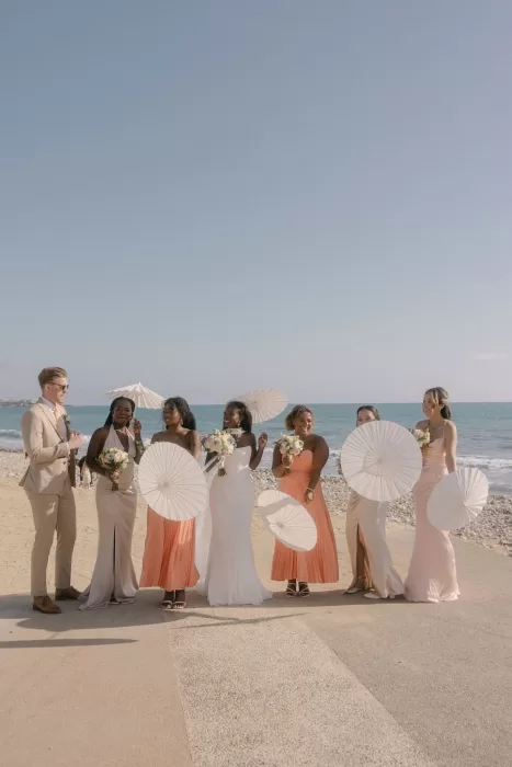 Bride with bridesmaids holding white parasols by the sea at Coral Residences.