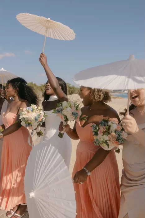 Bride and bridesmaids smiling and holding white parasols on the beach at Coral Residences.