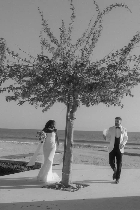Black and white photo of the couple smiling by a tree near the sea at Coral Residences.