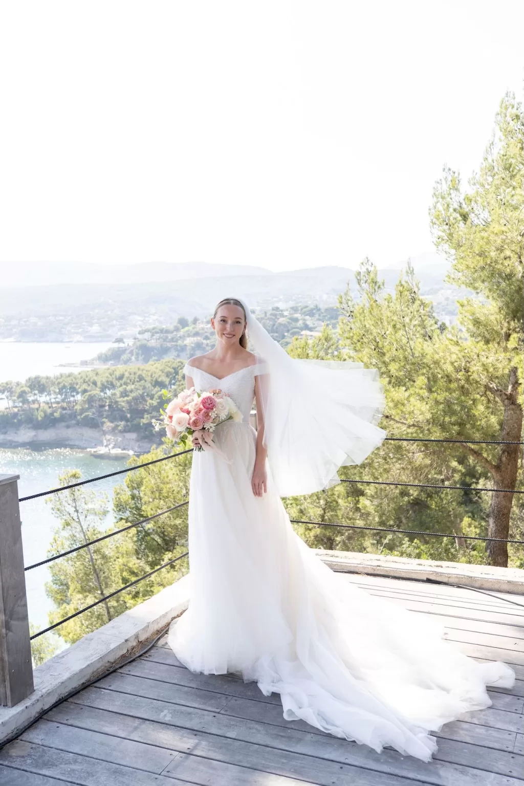 Bride in flowing gown holding a bouquet on a clifftop terrace at Domaine de Canaille, with sweeping sea and hillside views.