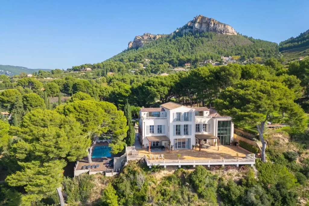 Aerial view of Domaine de Canaille, a white clifftop villa surrounded by lush greenery and mountain scenery.