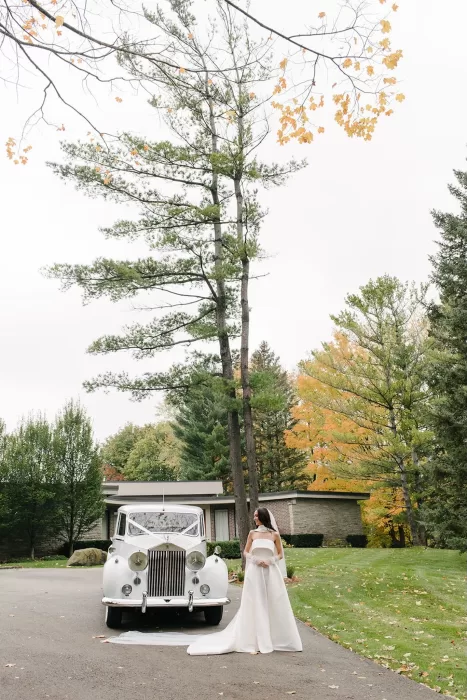 Bride standing beside a classic white car surrounded by autumn trees outside Graydon Hall.