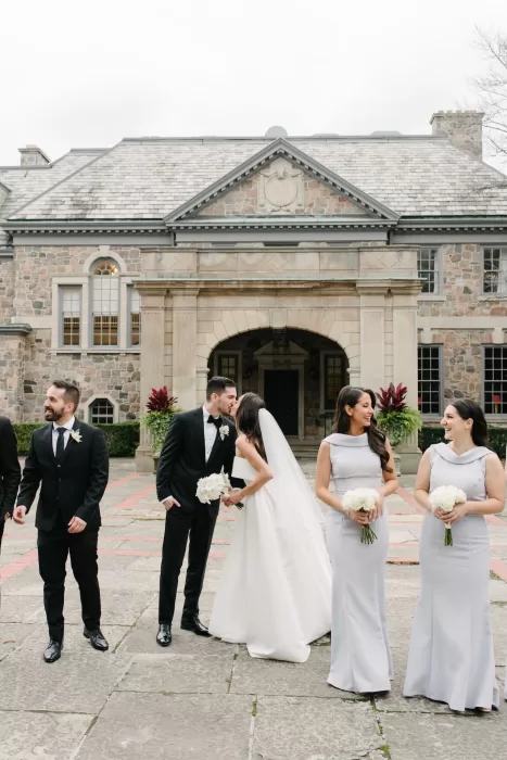 Wedding party smiling outside Graydon Hall as the bride and groom share a kiss in front of the grand entrance.
