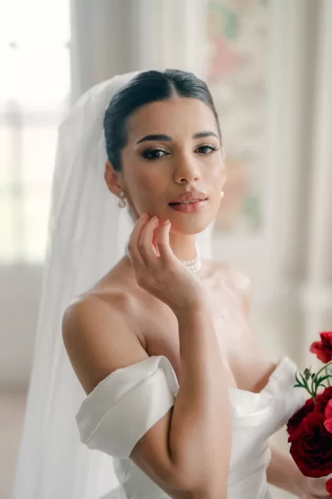 Close-up of bride touching her cheek, holding red bouquet, in soft light at Hedsor House.