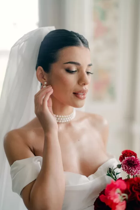 Bride adjusting pearl earrings with bouquet of red flowers at Hedsor House.