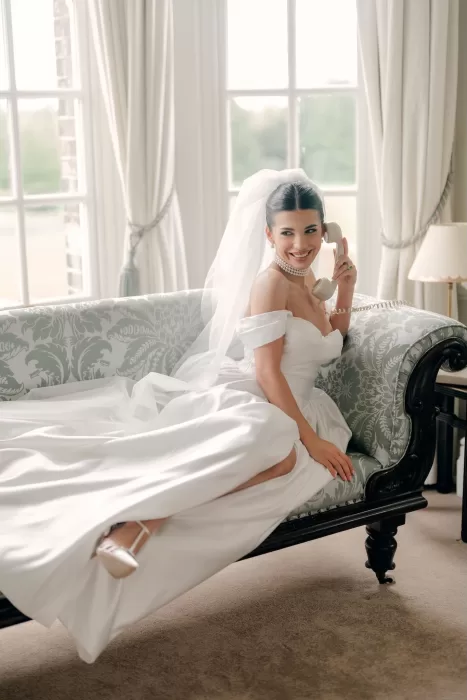 Bride in off-shoulder gown and pearl necklace smiling on a vintage sofa at Hedsor House.