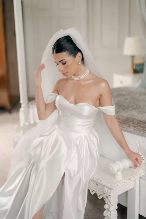 Bride in satin gown adjusting her veil while seated in a bridal suite at Hedsor House.