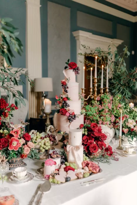 Elegant tiered wedding cake with red roses, grapes, and candles on a floral-decorated table at Hedsor House.