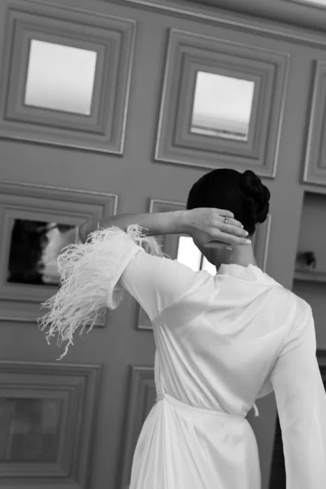 Black and white photo of the bride adjusting her hair in a feather-trimmed robe at La Farinera de Sant Lluis.