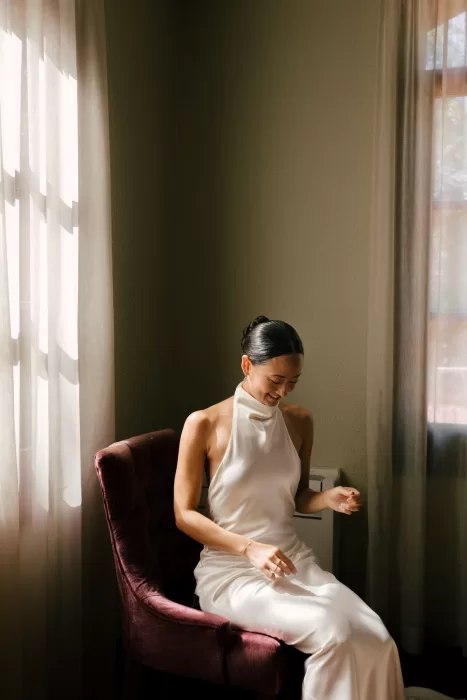 Bride smiling in soft window light while seated in a velvet chair before the wedding at La Farinera de Sant Lluis.
