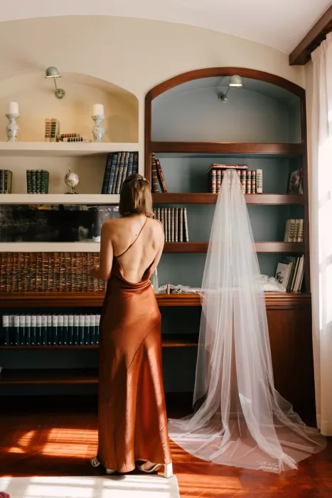 Bridesmaid in a rust satin gown standing beside bookshelves with the bridal veil hanging nearby at La Farinera de Sant Lluis.