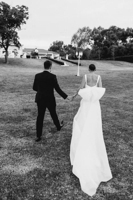 Bride and groom walking hand in hand across the lawn at Le Mas Barossa, her bow-back dress flowing behind her.