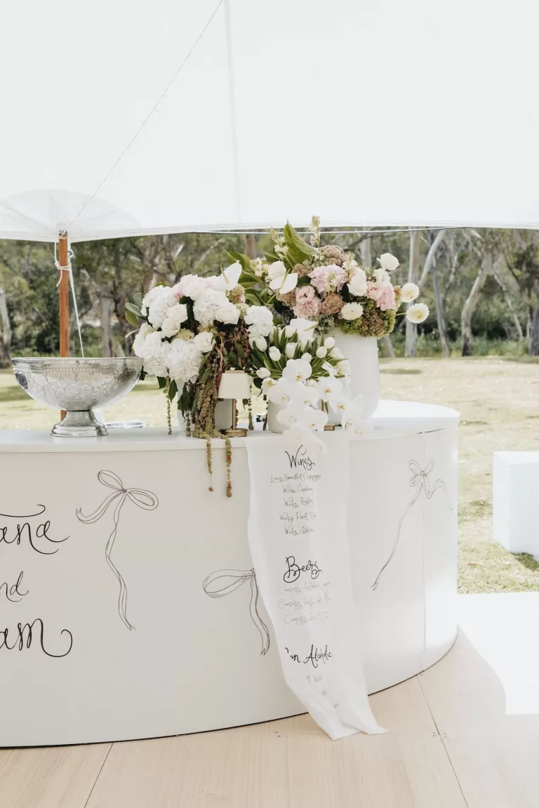 Curved white bar decorated with romantic floral arrangements and handwritten drinks menu at Le Mas Barossa.