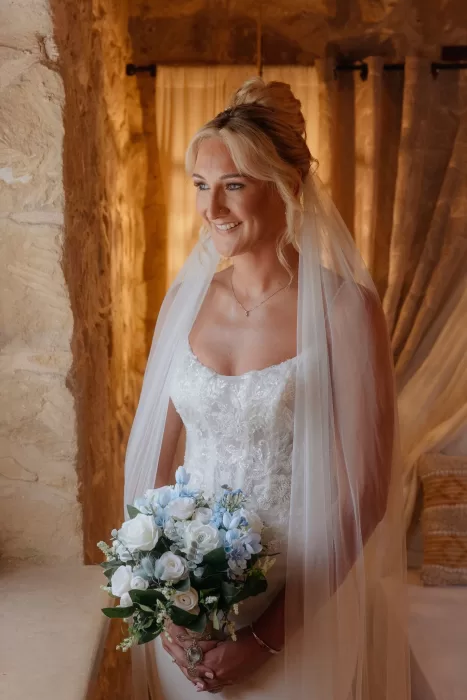 Bride holding a bouquet of white and blue flowers, smiling softly inside Liopetro’s stone interior.