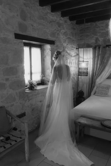 Bride in her wedding gown standing by a rustic stone window at Liopetro, veil flowing behind.