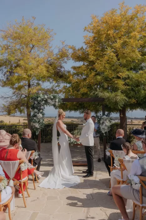 Bride and groom exchanging vows under a floral arch at Liopetro with guests seated around them.