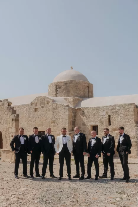 Groom with his groomsmen in tuxedos posing outside the stone chapel at Liopetro.