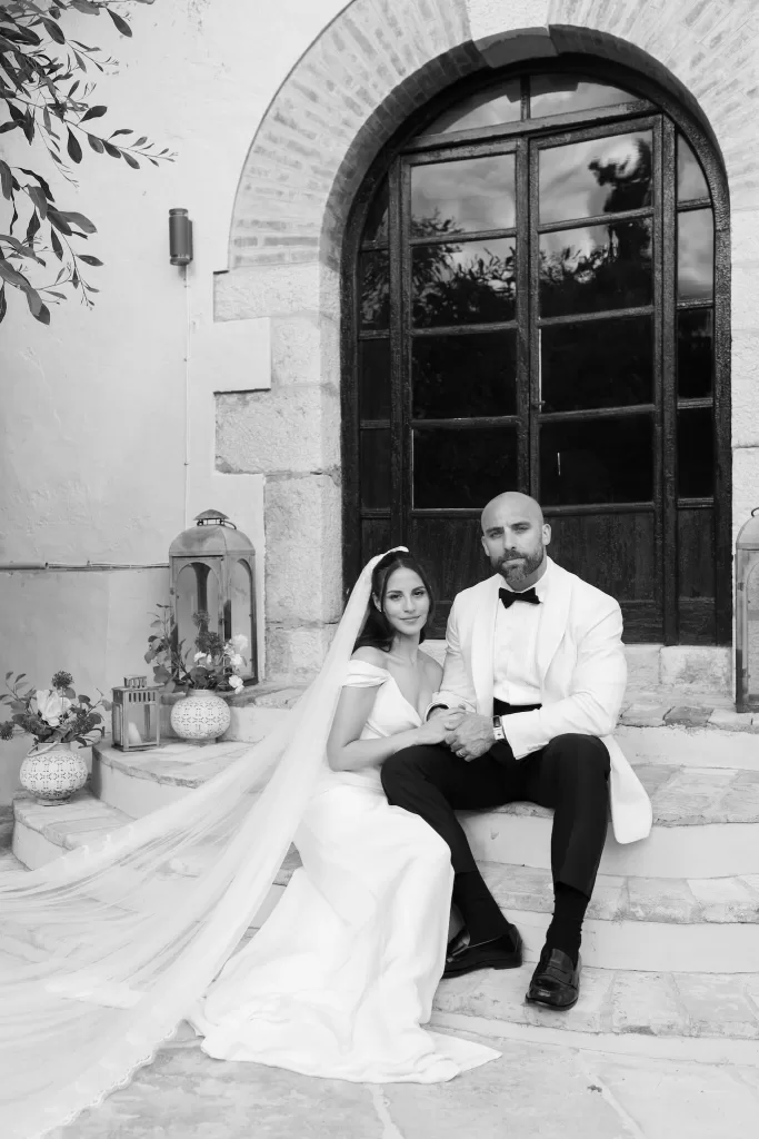 Black and white image of bride and groom seated on steps outside Masia Victoria.
