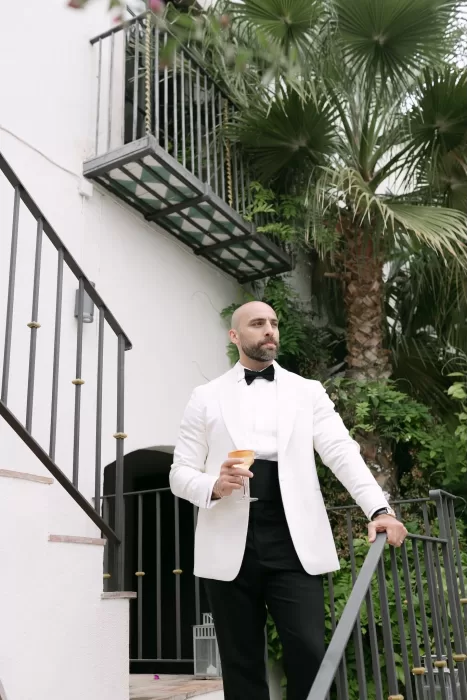 Stylish groom in a white tuxedo holding a drink on the staircase at Masia Victoria.
