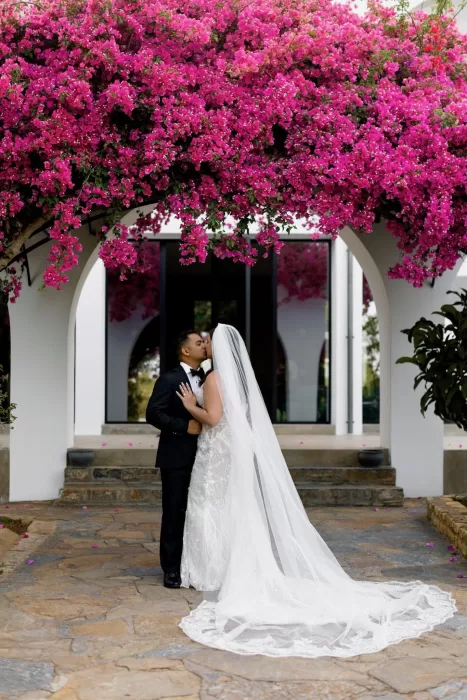 Bride and groom kissing under vibrant pink bougainvillea arch at Minos Beach Art Hotel.