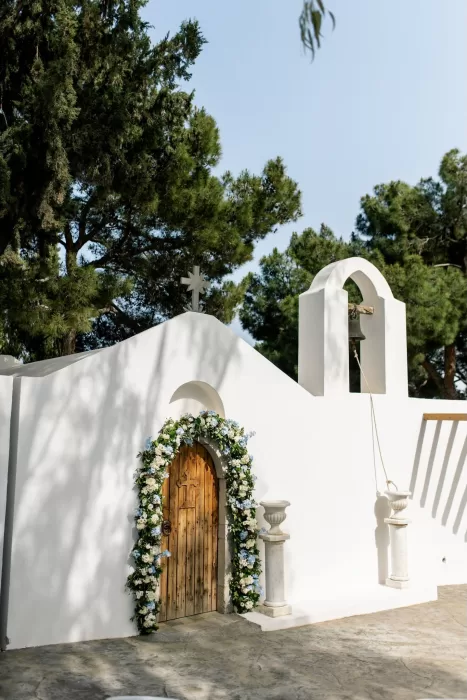 Whitewashed chapel with wooden door framed by blue and white floral arch at Minos Beach Art Hotel.