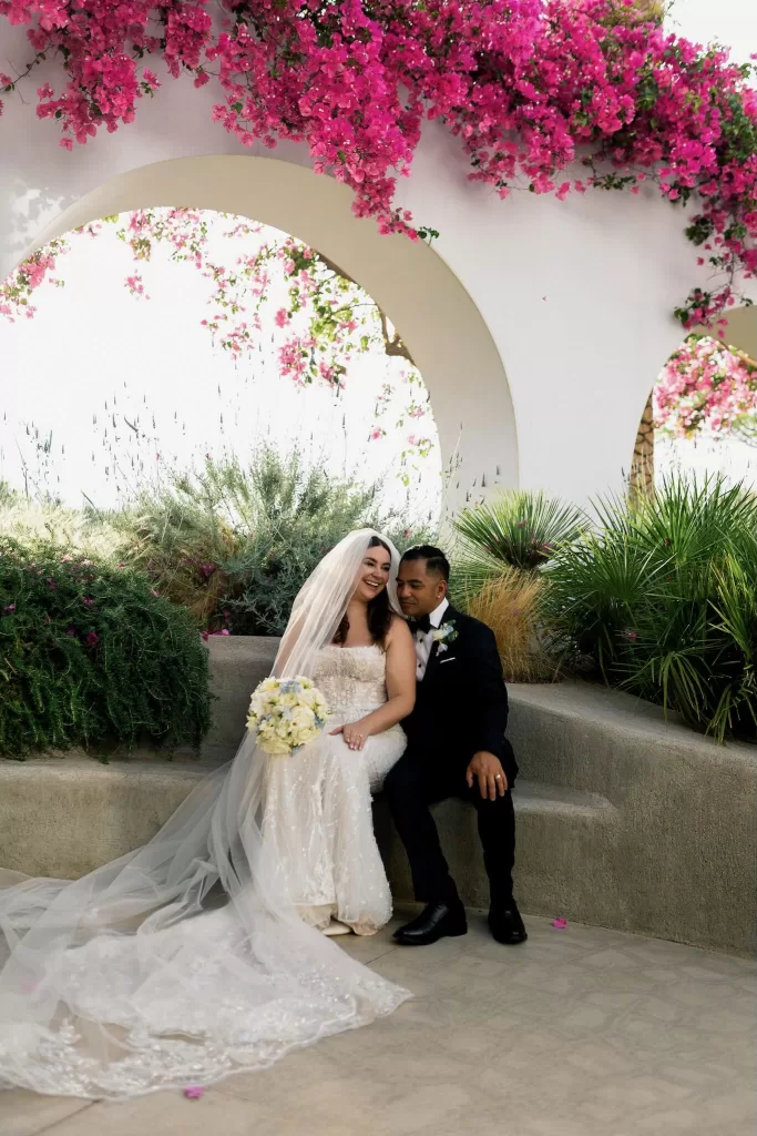 Bride and groom seated under white arch at Minos Beach Art Hotel, surrounded by lush greenery and vibrant pink bougainvillea.