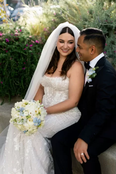 Bride and groom sitting together, smiling, with a bouquet of white and blue flowers at Minos Beach Art Hotel.