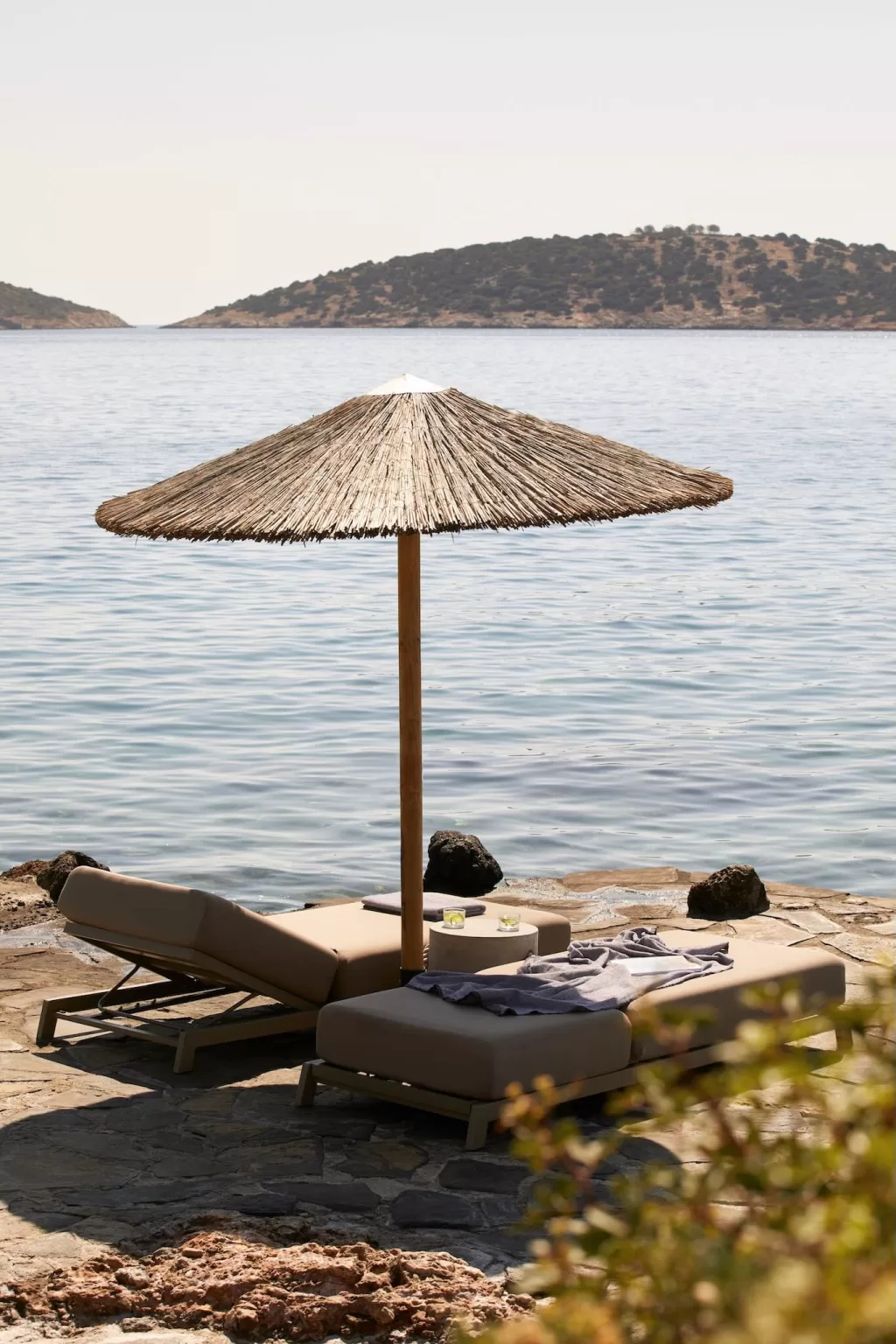 Seafront loungers with a thatched umbrella at Minos Beach Art Hotel overlooking calm blue waters and distant islands.