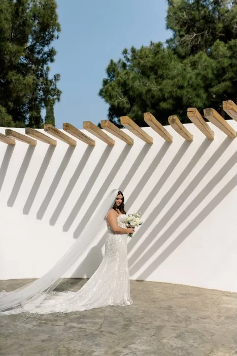 Bride holding bouquet beneath striking shadows cast on white wall at Minos Beach Art Hotel.