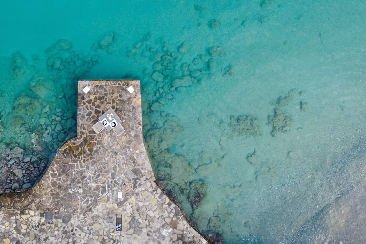 Aerial view of a stone pier with dining table over turquoise waters at Minos Beach Art Hotel.