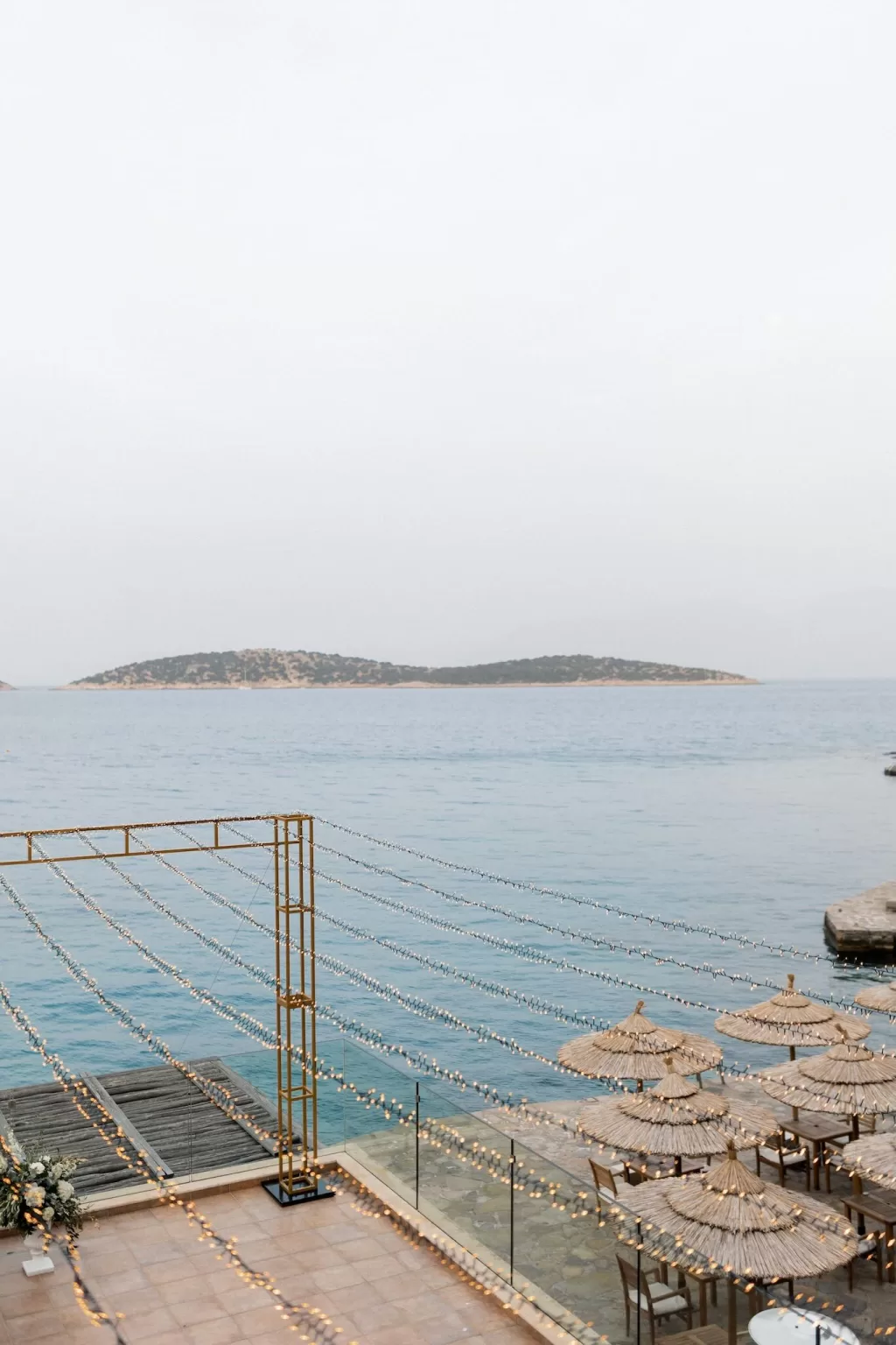 View of oceanfront dining area with string lights and straw parasols at Minos Beach Art Hotel.