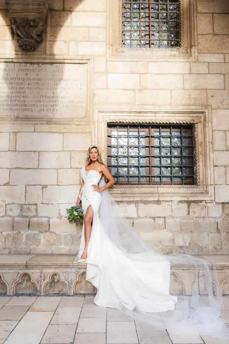 Bride in an elegant white gown with a long train posing against stone walls at the Museum of Modern Art Dubrovnik.