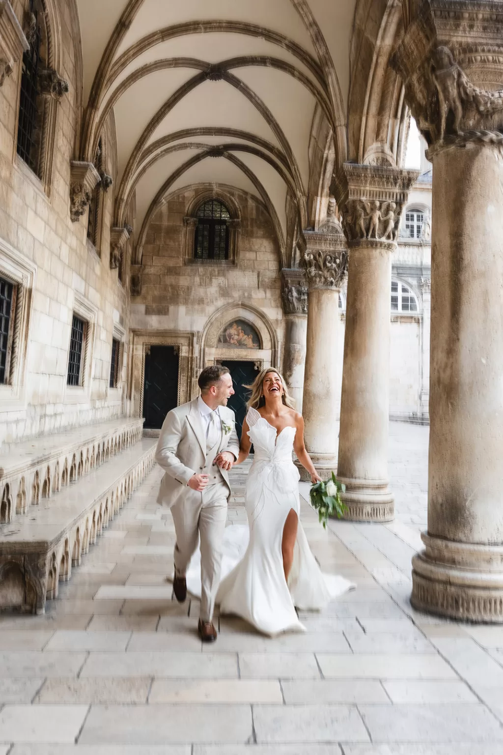 Bride and groom walking hand in hand outside the stone arches of Museum of Modern Art Dubrovnik at night.