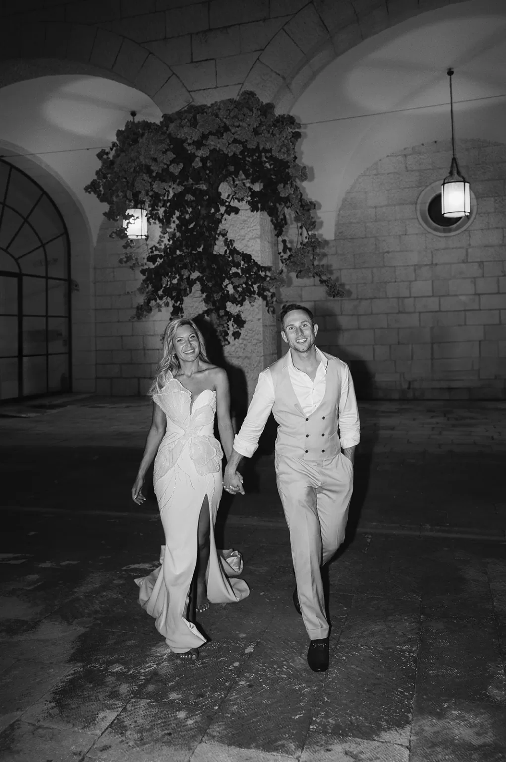 Bride and groom walking hand in hand outside the stone arches of Museum of Modern Art Dubrovnik at night.