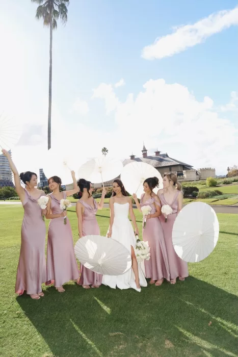 Bride surrounded by bridesmaids in blush satin dresses holding white parasols on the green lawn of Next Chapter Events.