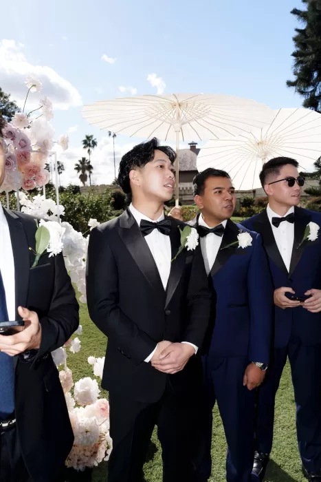 Groomsmen in black and navy suits holding white parasols during the sunny outdoor ceremony at Next Chapter Events.