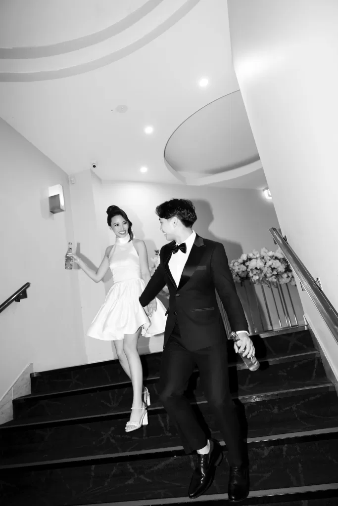 Bride in a short reception dress and groom descending stairs hand in hand, smiling and holding drinks at Next Chapter Events.