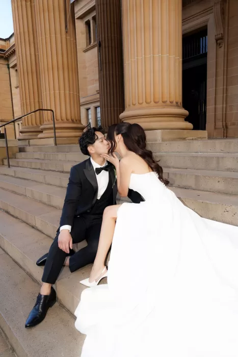 Bride and groom sharing a kiss on grand sandstone steps outside Next Chapter Events, her gown flowing elegantly around them.