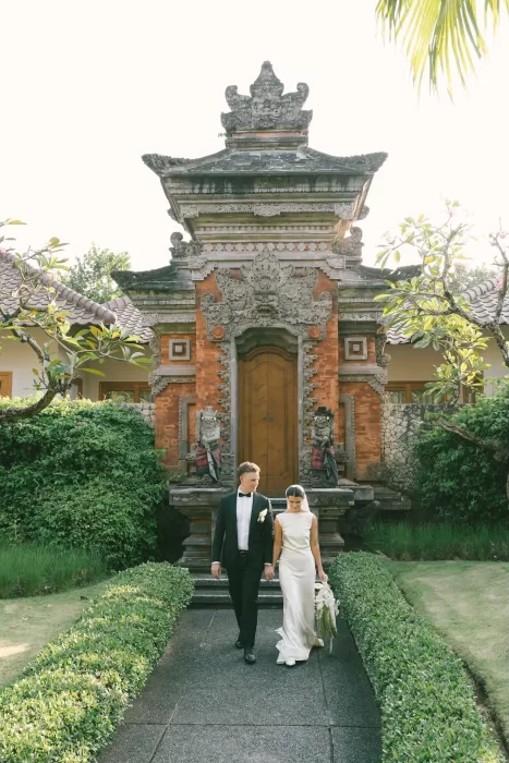 Bride and groom walking hand in hand past a traditional Balinese gate at Padma Resort Legian.