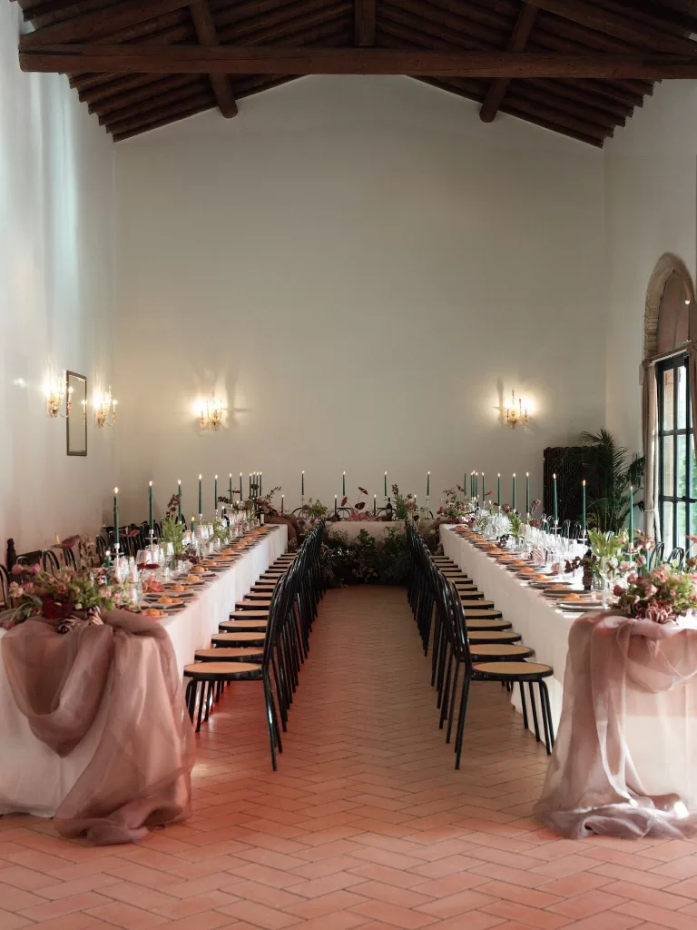 Long banquet tables with floral arrangements and candles set in the rustic hall of Rustico Bertel.