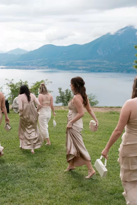 Bridesmaids in satin dresses walking across the lawn with a sweeping lake and mountain view at Rustico Bertel.