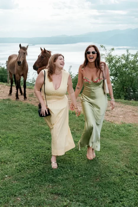 Two wedding guests laughing while walking outside at Rustico Bertel with horses and lake views in the background.